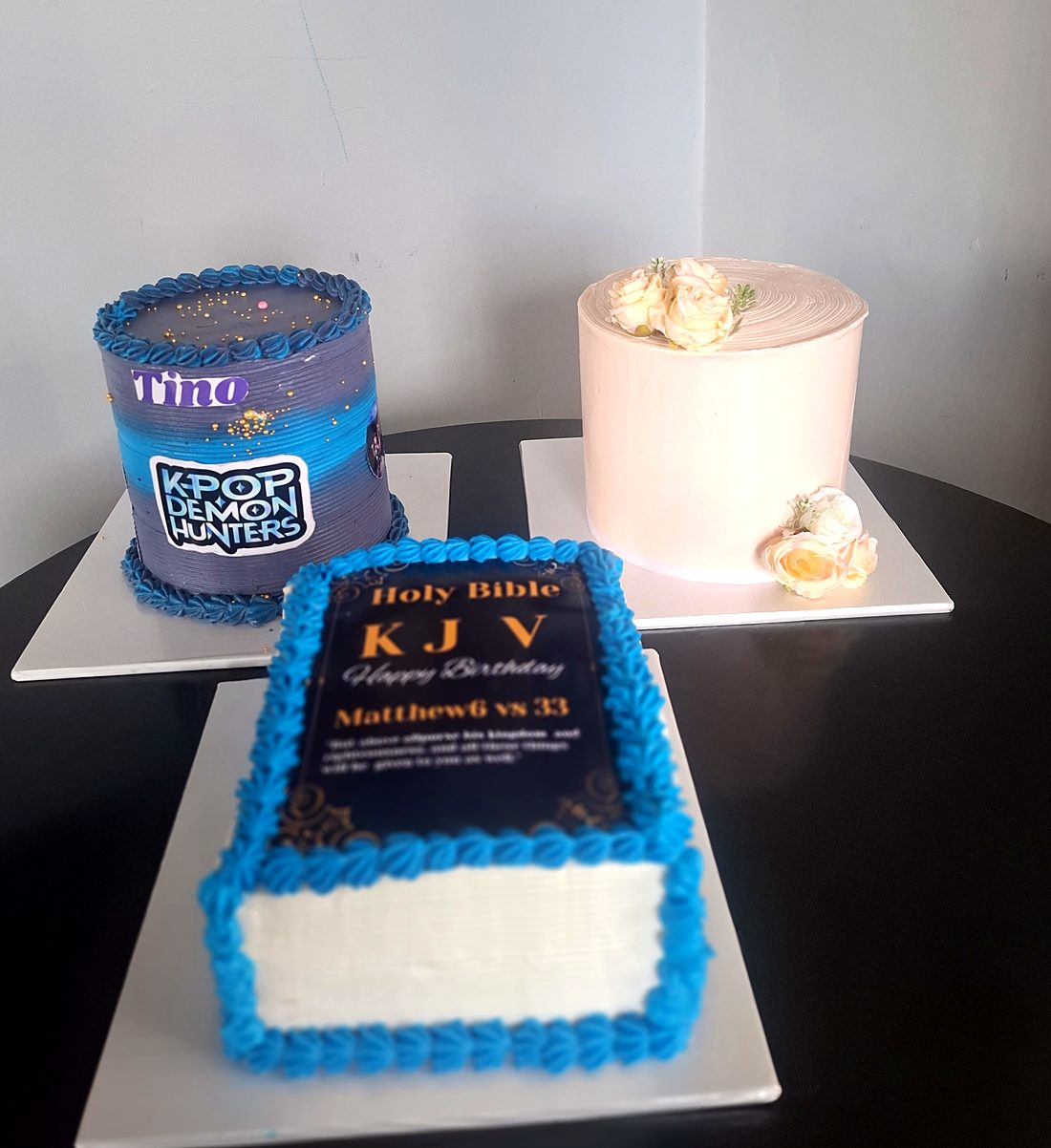 Three birthday cakes: a Holy Bible book cake, a K-Pop Demon Hunters cake and a blush pink cake with sugar flowers